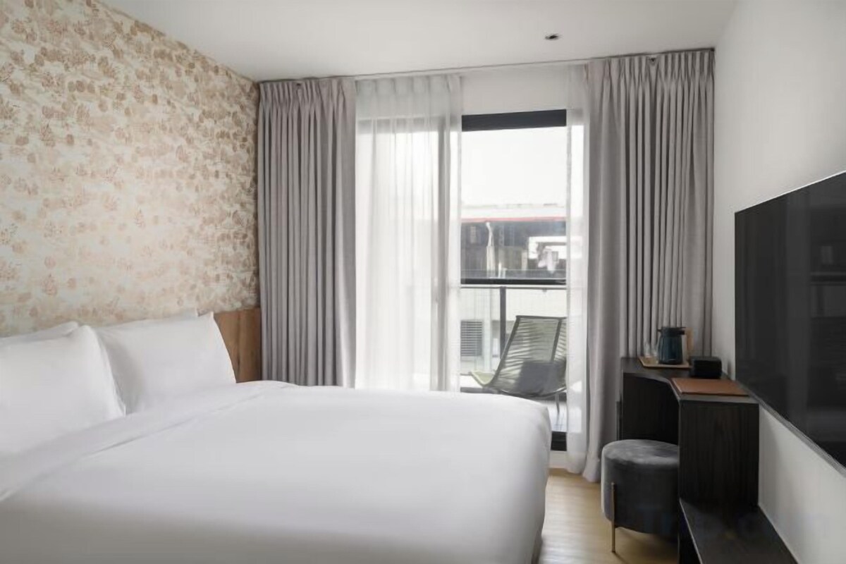 A hotel room with a large bed, a TV, a desk, and a view of the city through the window.