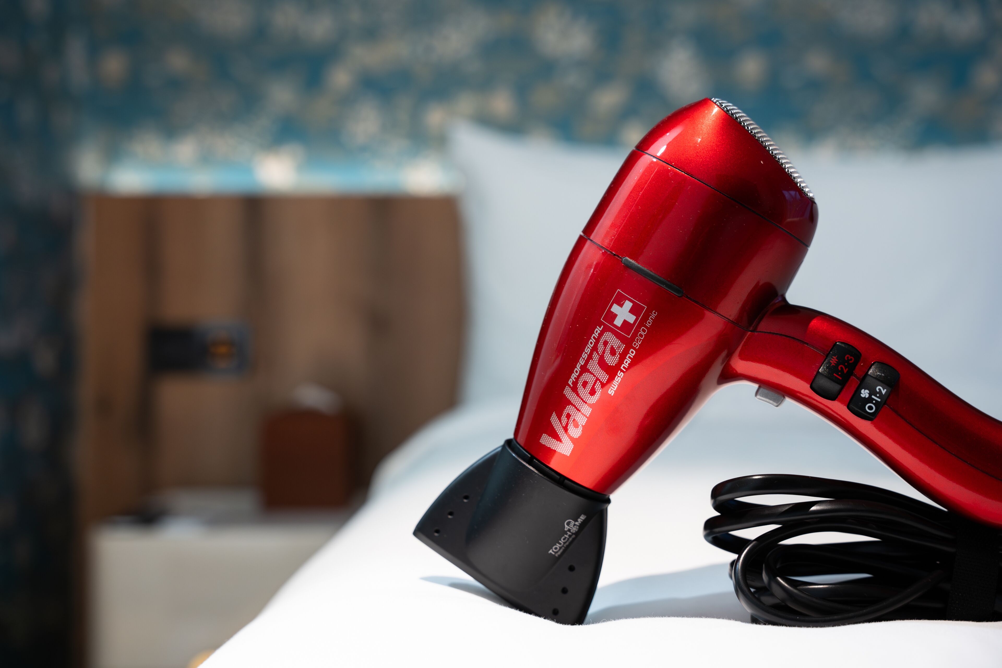 A red hair dryer with a black base and a Swiss flag logo, placed on a white surface with a blurred background.
