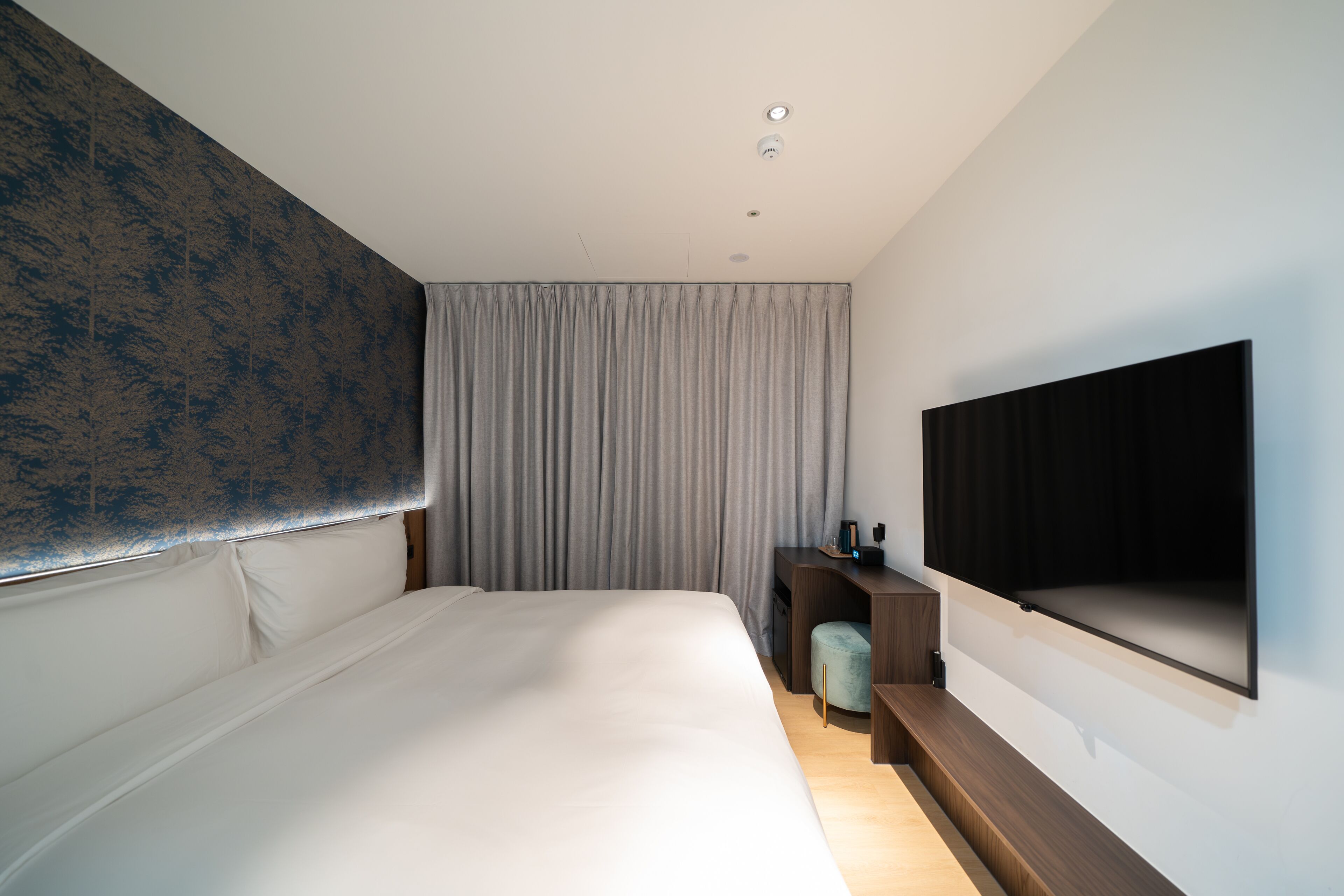Ein modernes Hotelzimmer mit einem großen Bett, einem Schreibtisch mit Stuhl, einem flachbildfernseher an der Wand und einem großen Fenster mit Vorhängen.
