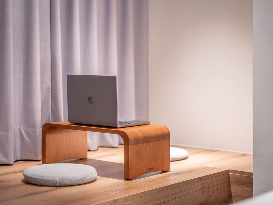 Ein Laptop auf einem Holzständer mit Kissen, eine hölzerne Bank und ein Kissen am Boden.