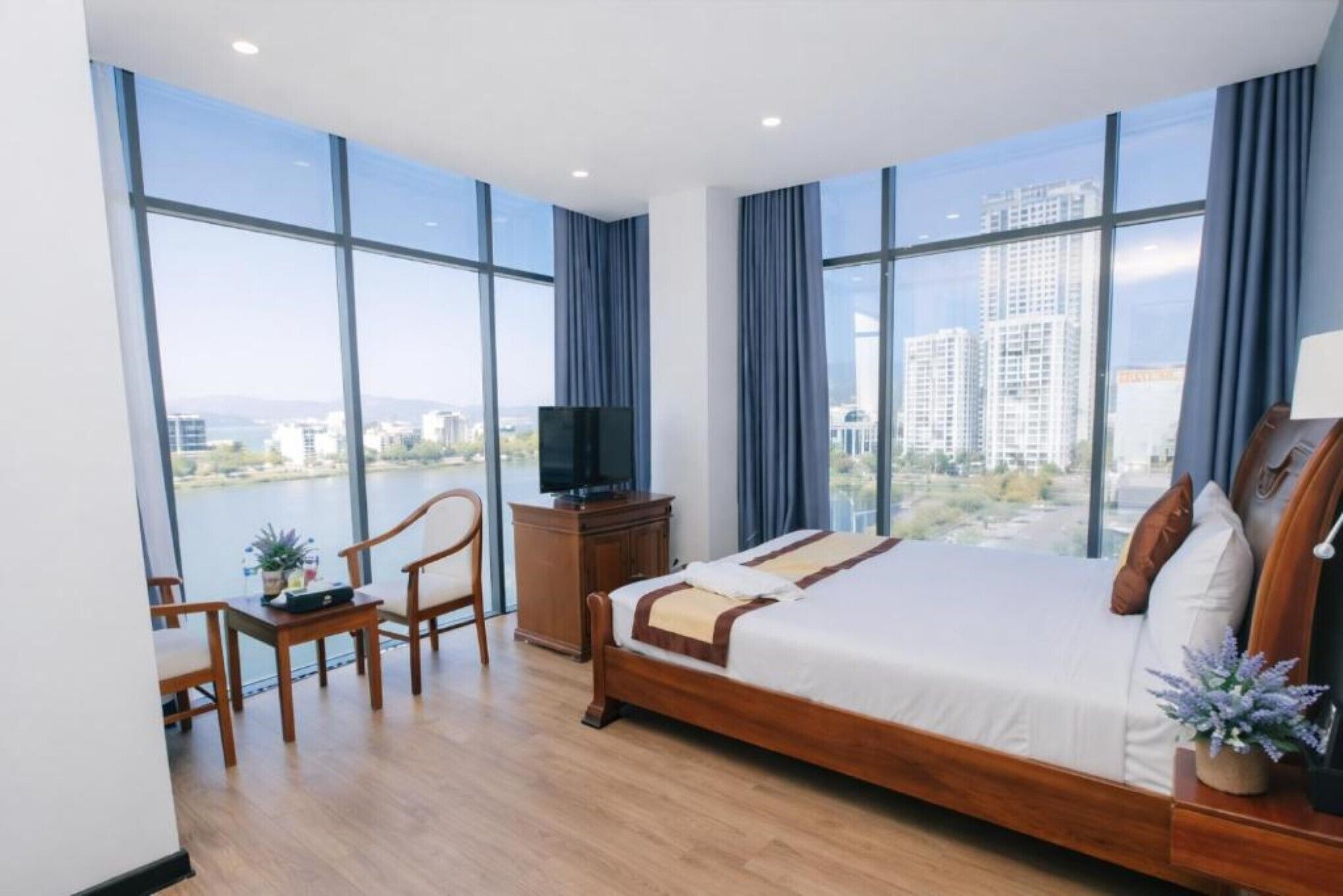 Habitación de hotel moderna con una cama grande, televisión, escritorio y vistas a un paisaje urbano a través de amplios ventanales.