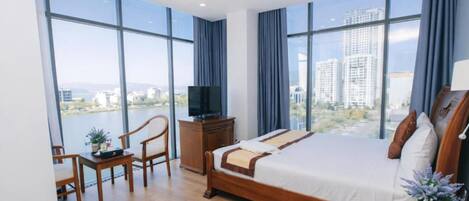 Habitación de hotel moderna con una cama grande, televisión, escritorio y vistas a un paisaje urbano a través de amplios ventanales.