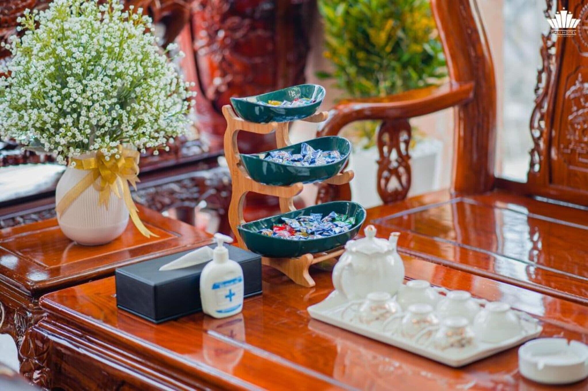 A wooden table with a vase of white flowers, a bowl of snacks, a bottle of hand sanitizer, and a tray of condiments.