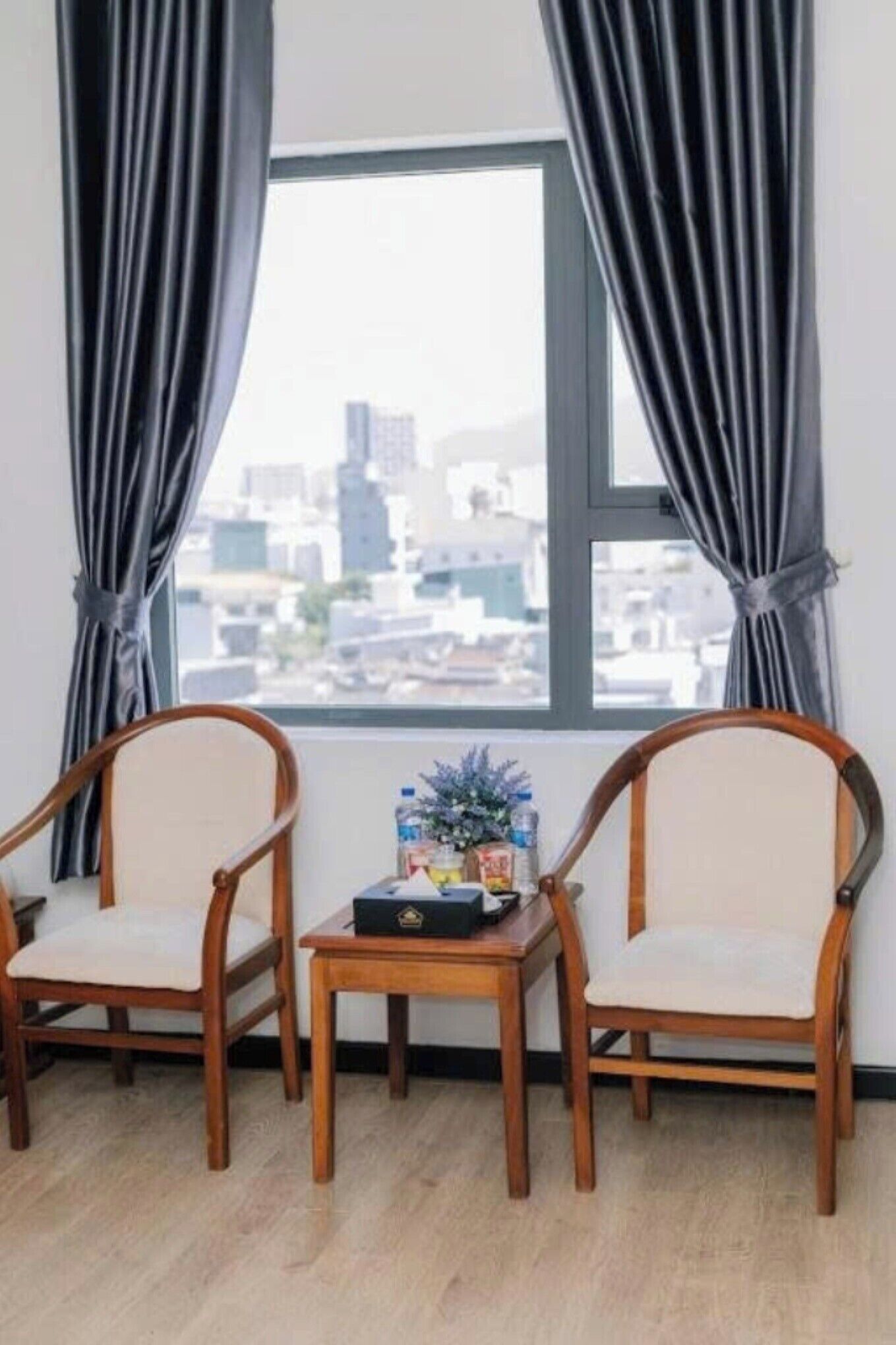 Um quarto com duas cadeiras de madeira, uma mesinha com uma garrafa e uma caixa, e uma janela com cortinas.
