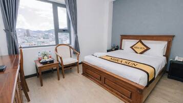 A hotel room with a bed, a desk, a chair, a nightstand, and a view of the city through large windows.