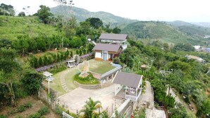 A large house with a spacious patio, surrounded by lush greenery and hills in the background.