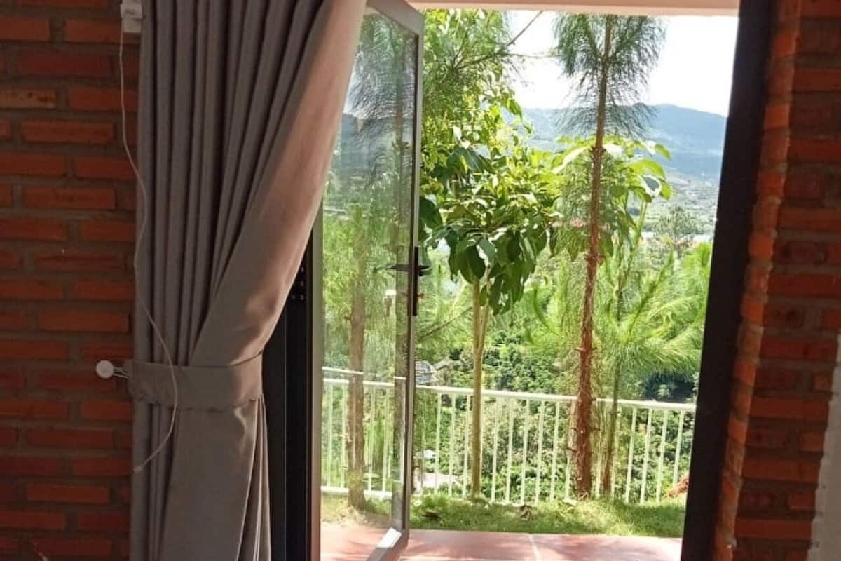 Ein Zimmer mit einem Bett, einem kleinen Tisch und einer Glasschiebetür, die auf einen Balkon mit Blick auf Grünflächen und einen Zaun führt.