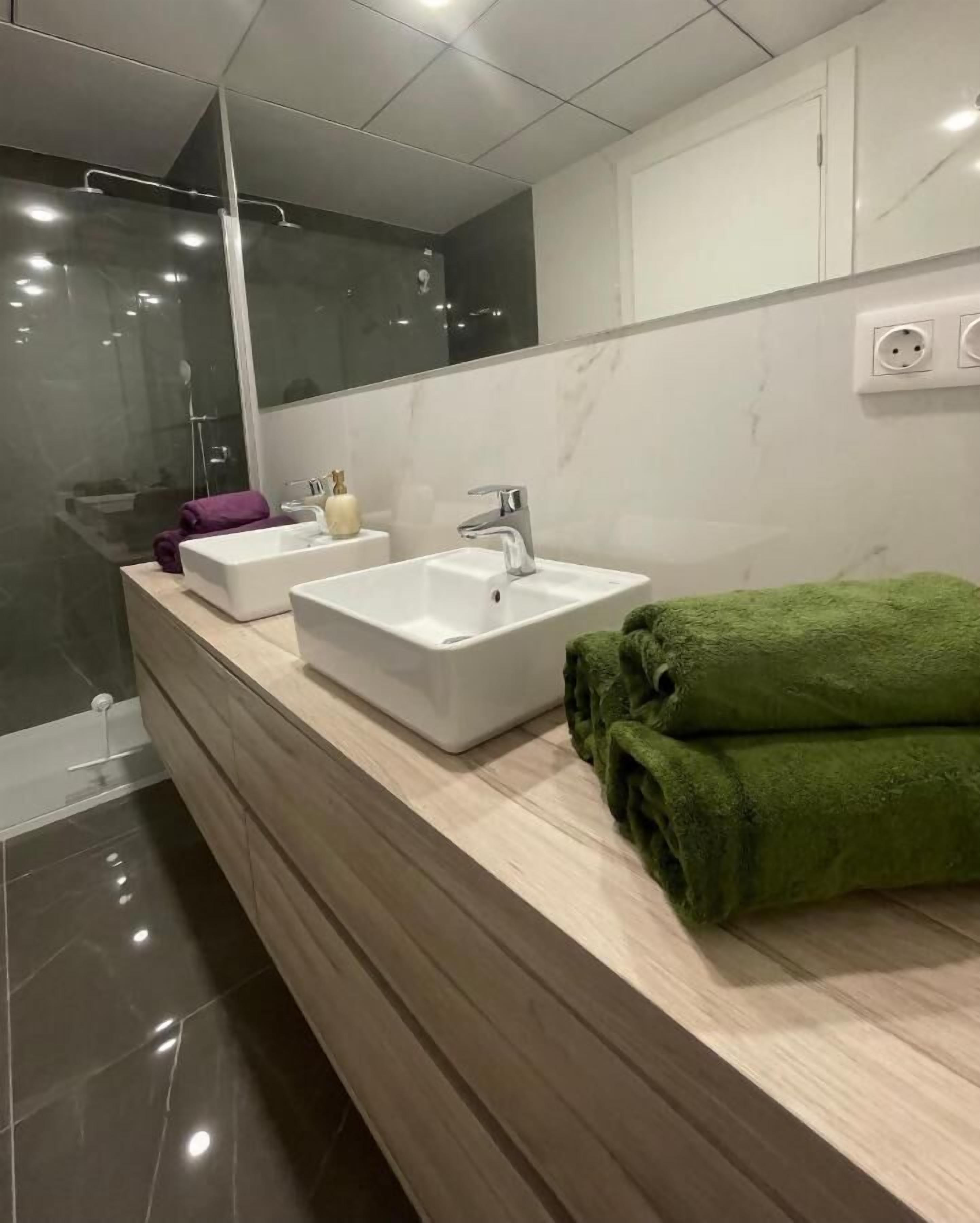 Ein modernes Badezimmer mit Duschkabine aus Glas, zwei Waschbecken, einem Spiegel und einem grünen Handtuch auf der Ablagefläche.
