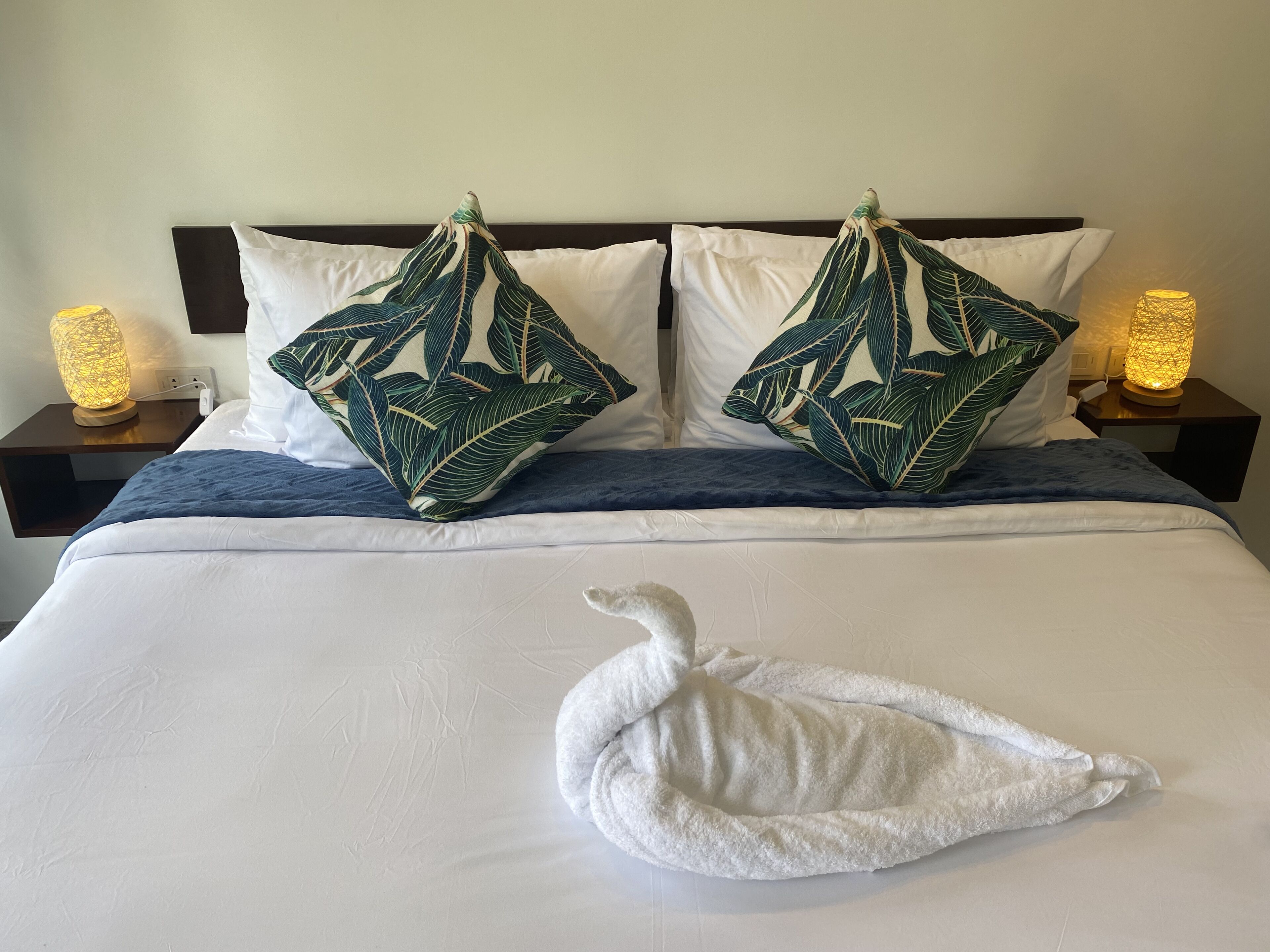 Un lit bien fait, avec du linge de lit blanc, deux coussins décoratifs ornés d’un motif de feuilles et une serviette blanche pliée posée sur le lit.