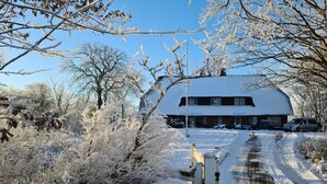 A snow-covered rural road leading to a house with a thatched roof and a car parked outside.