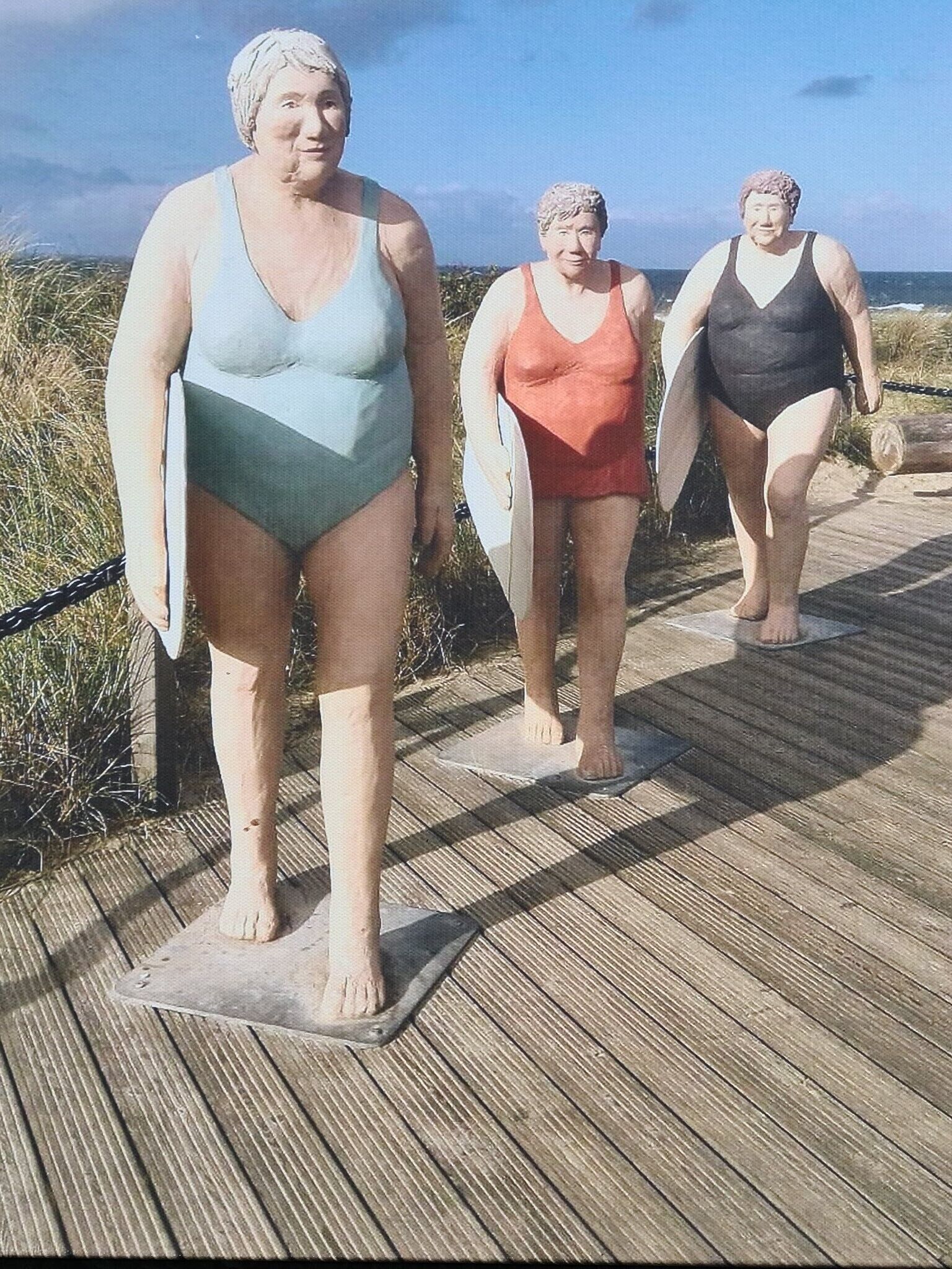 A wooden boardwalk with three statues of people in swimsuits.