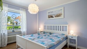 Une chambre à coucher comprenant un lit, des tables de chevet, un luminaire suspendu, une fenêtre avec des rideaux et un tableau accroché au mur.