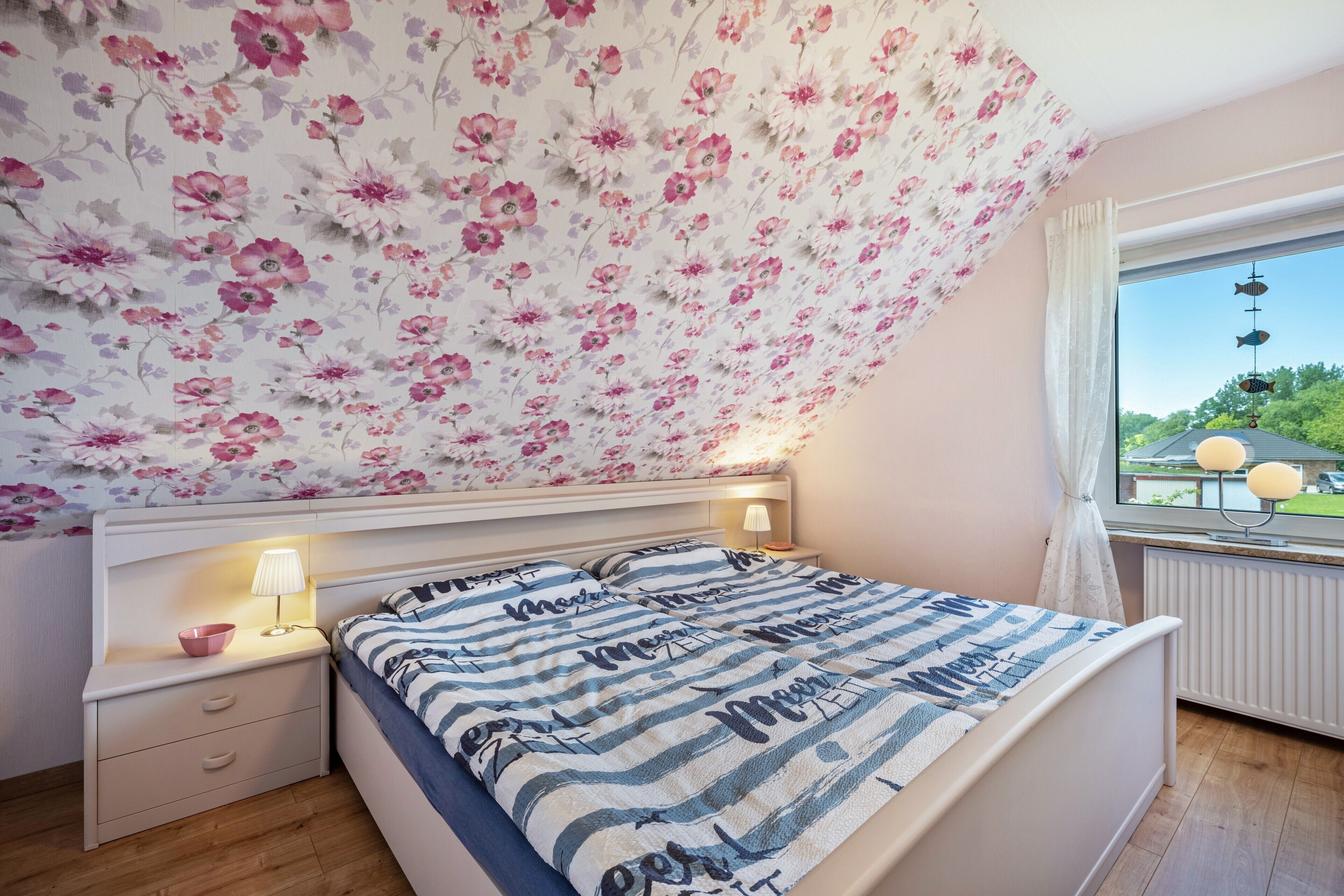 Makuuhuone, jossa on kalteva katto, raidallinen vuodevaatteilla varustettu sänky, yöpöytä lampun kanssa ja ikkuna, josta avautuu näkymä vehreään maisemaan.