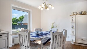 Un coin repas avec une table dressée pour quatre personnes, des chaises blanches, un lustre et une vue sur un bâtiment à l’extérieur.