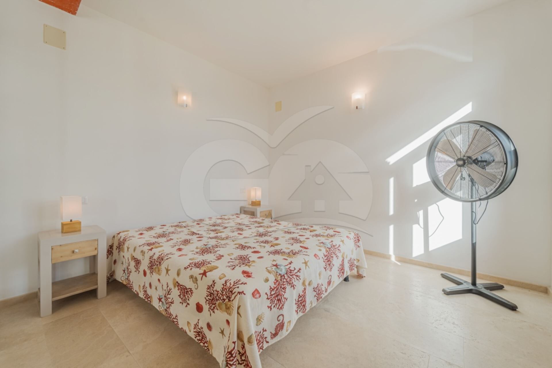 A bedroom with a bed, bedside tables, a standing fan, and a window.