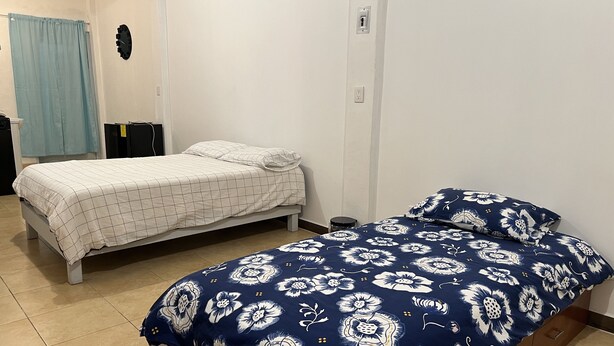 A bedroom with two beds, one with a floral bedspread and the other with a checkered bedspread. There is a small bedside table and a wall clock. - Apartment 32mt2 Tultitlán Edomex. Near the municipal palace. Beautiful garden  (Tultitlán de Mariano Escobedo)