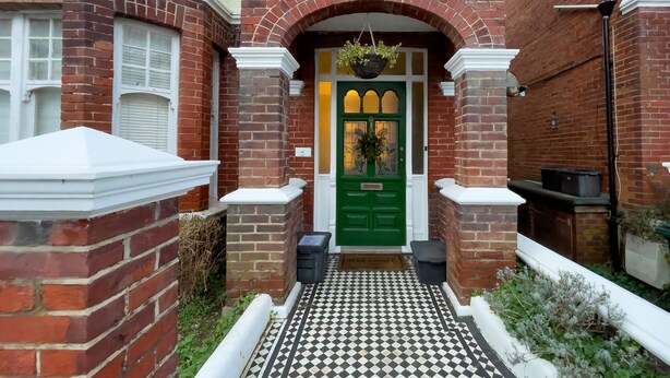 Una casa de ladrillo rojo con puerta verde, cerca blanca enrejada y piso a cuadros blanco y negro.