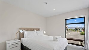Habitación de hotel con cama, mesita de noche, lámpara y un gran ventanal con vista a un estacionamiento y edificios.