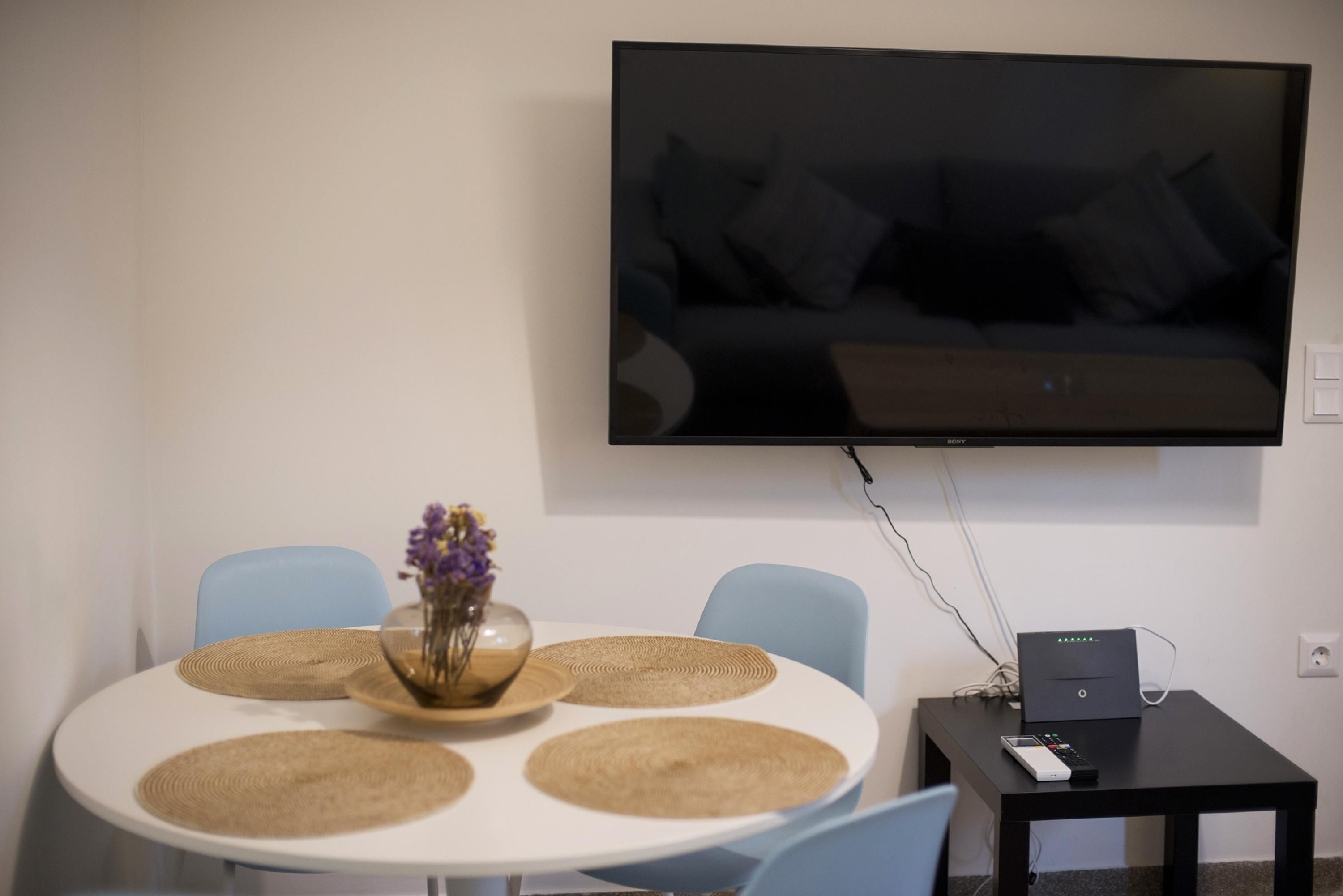 Un salon moderne avec un téléviseur à écran plat fixé au mur, une table à manger ronde avec des sets de table, un vase rempli de fleurs et une petite table noire sur laquelle repose une télécommande.
