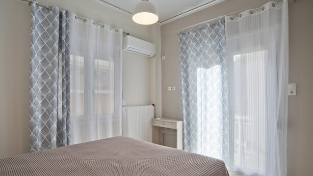 Ein Schlafzimmer mit Bett, Nachttisch, Fenster mit leichtem Vorhang und Klimaanlage.