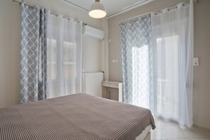 A bedroom with a bed, a bedside table, a window with sheer curtains, and an air conditioning unit. - Piraeus Wave in Piraeus (Piraeus)