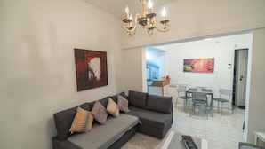 A modern living room with a sectional sofa, a chandelier, and artwork on the walls. - Marina Apartment in Nikea (Nikaia-Agios Ioannis Rentis)