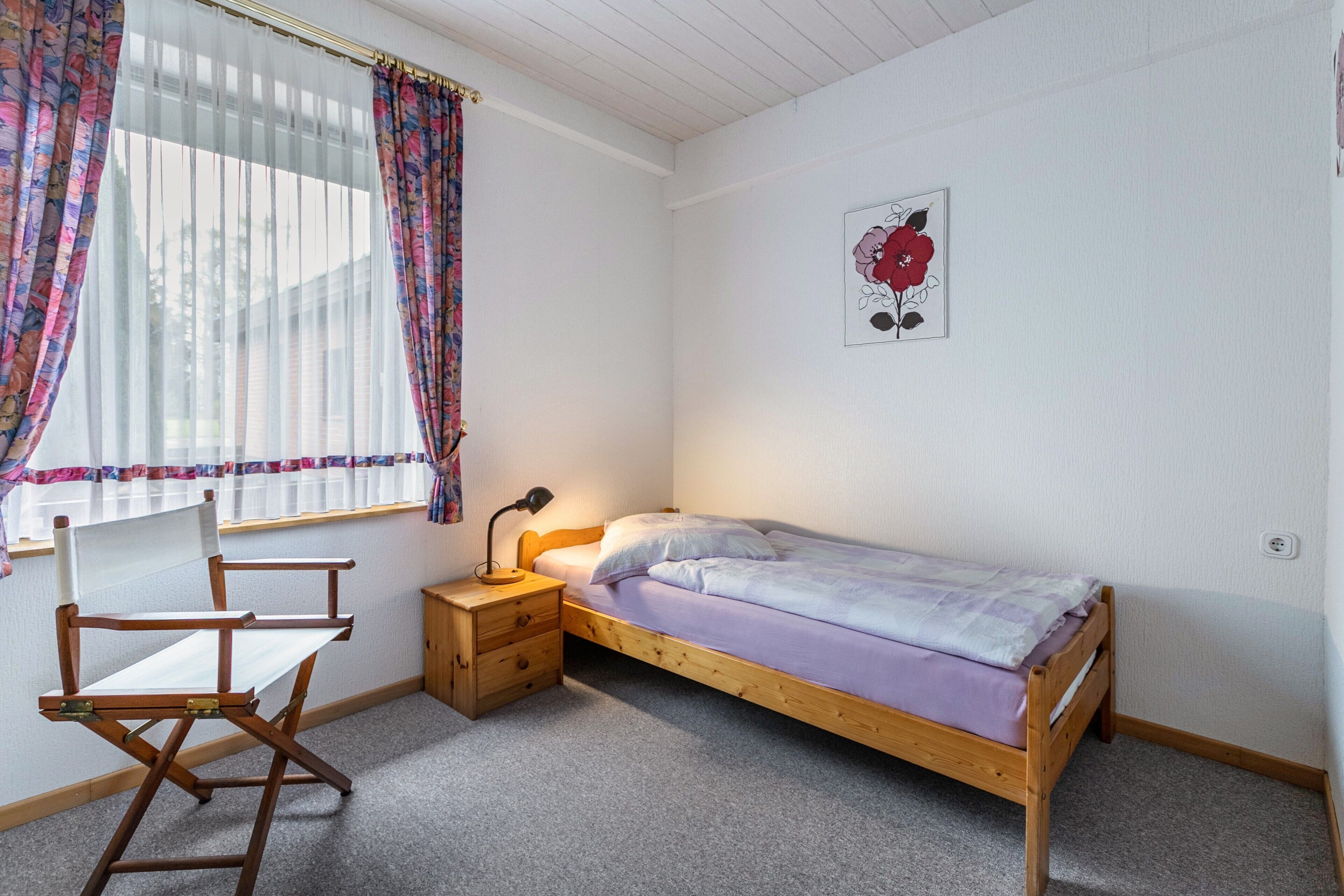 A single bed with a wooden frame, a small bedside table with a lamp, a chair, and a window with sheer curtains.
