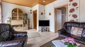 A living room with a wooden ceiling, a television, leather sofas, a bookshelf, and a floral wall decoration.