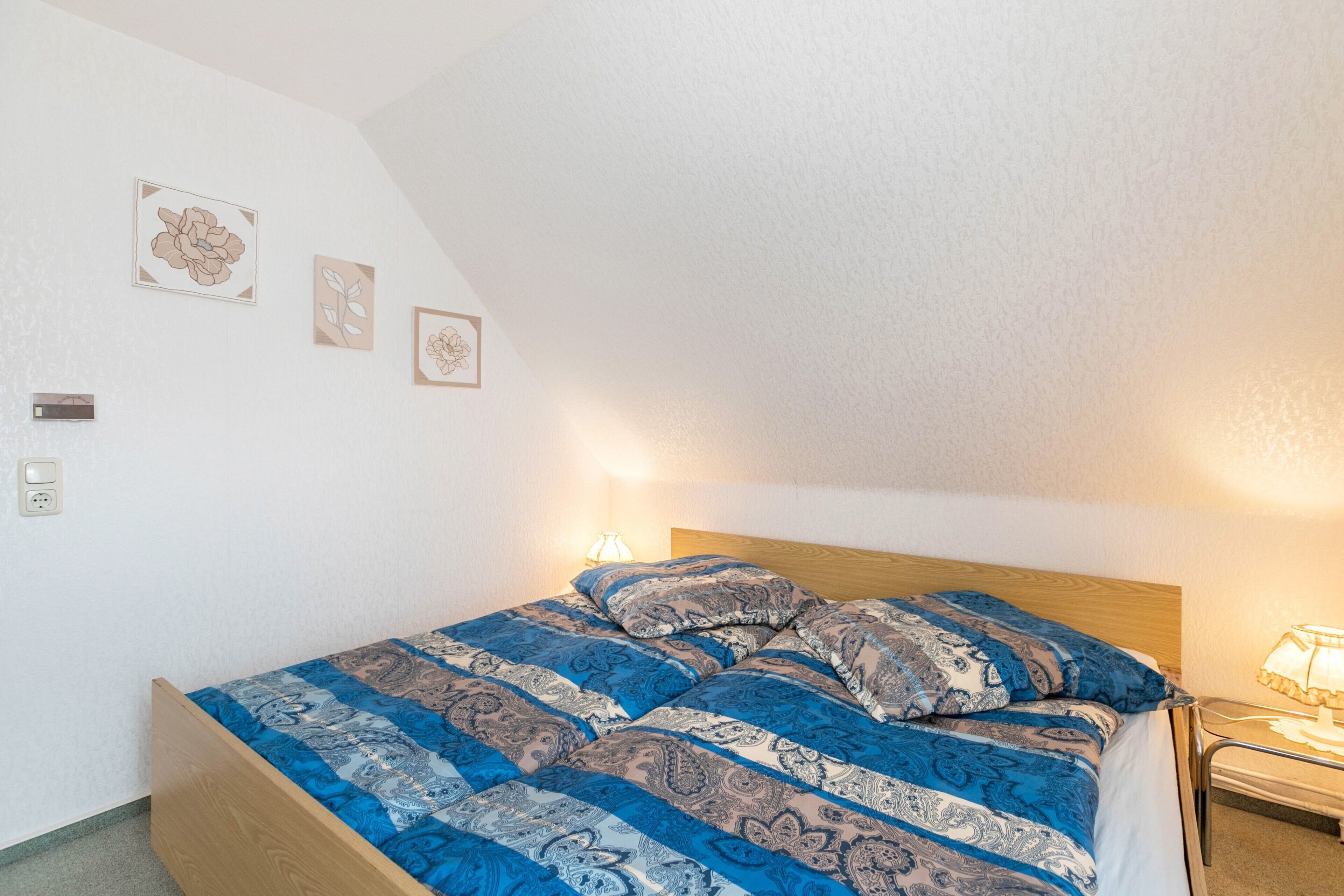A bedroom with a wooden bed, patterned bedding, a nightstand with a lamp, and wall-mounted artwork.