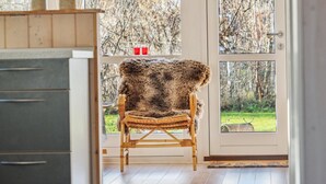 Ein Zimmer mit einem mit Fell bezogenen Holzstuhl, einer Küchenzeile mit Schränken und Blick auf Bäume durch eine Glastür.