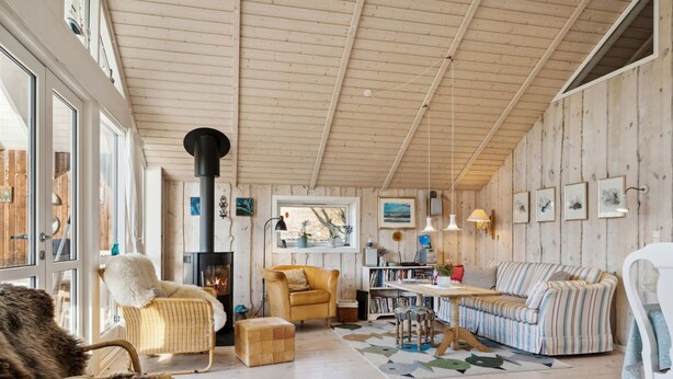 Ein gemütliches Blockhaus-Interieur mit Holzbalkendecke, Kamin, gestreifter Couch, Korbsessel und Esstisch.