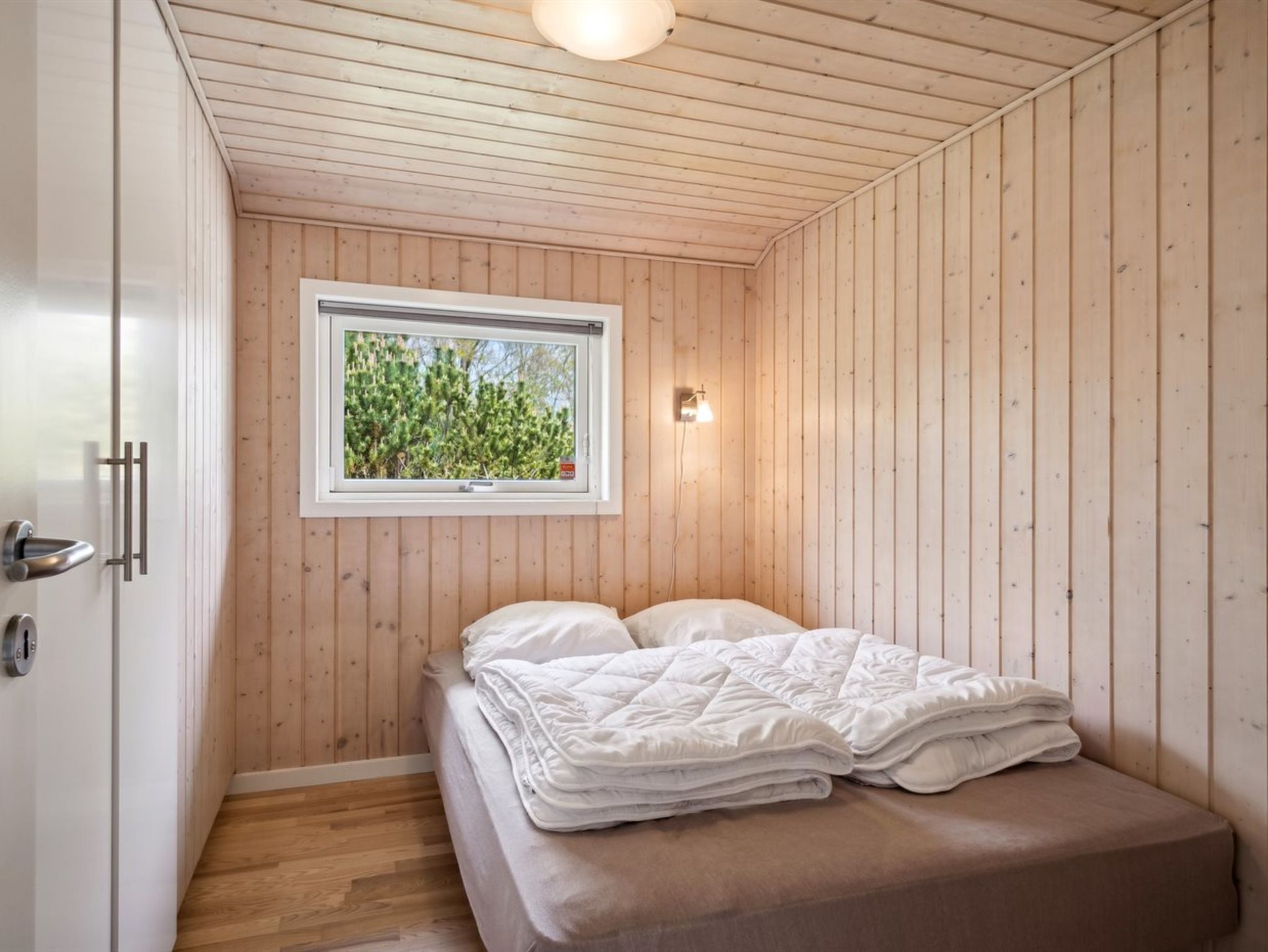 A small, cozy bedroom with wooden walls, a single bed, a window with a view of greenery, and a wall-mounted light fixture.