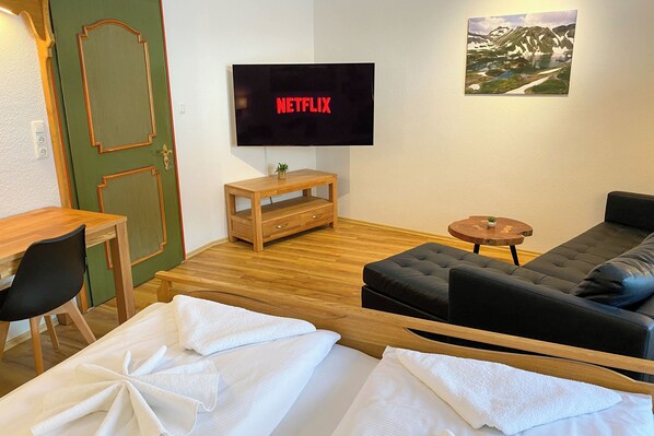 A modern hotel room with a wooden floor, a black sofa, a wooden desk, a television displaying 'NETFLIX', and a mountain landscape painting on the wall. - Haus Breitenberg (Bad Hindelang)