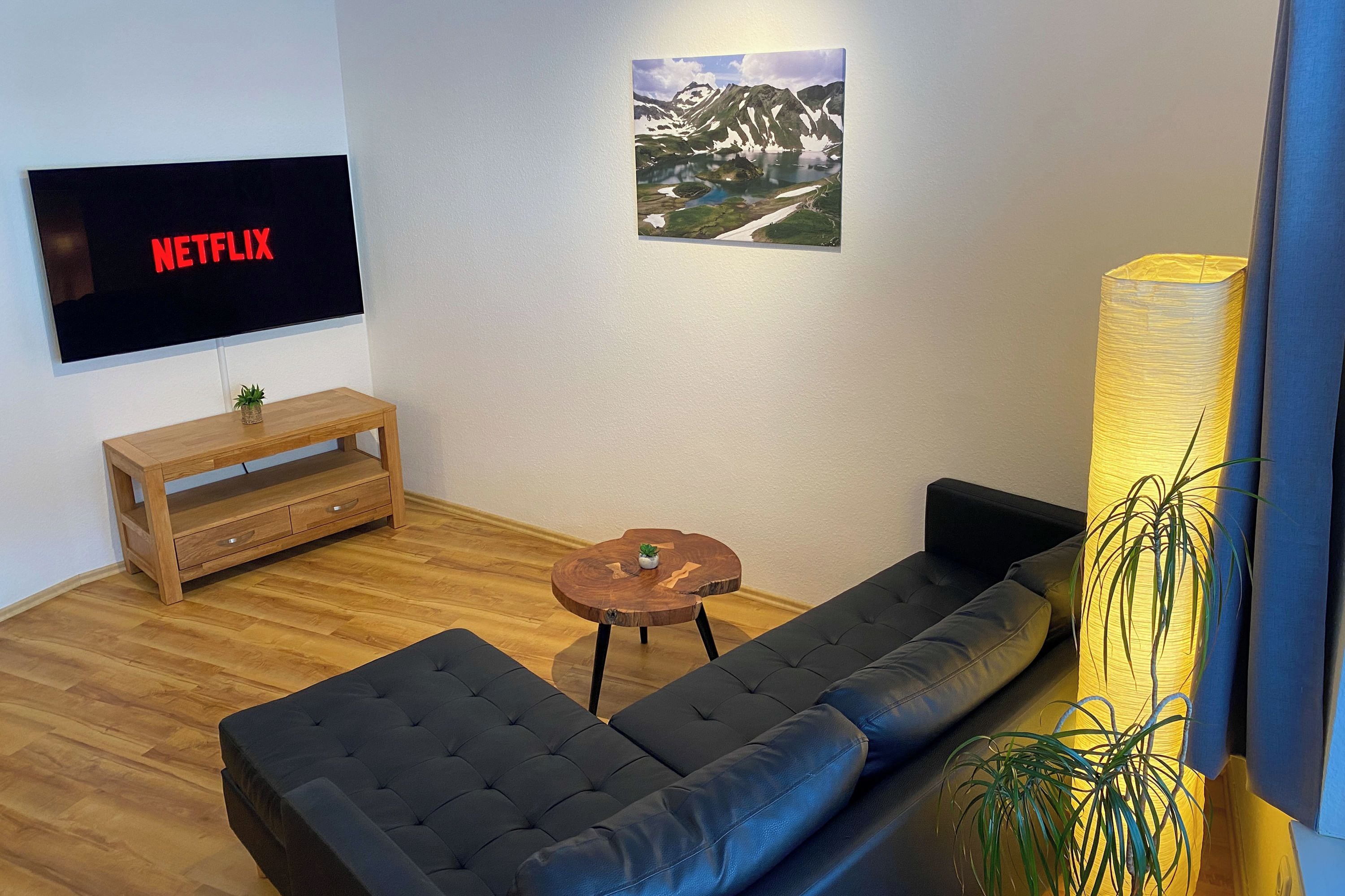 A modern living room with a large flat-screen TV displaying 'NETFLIX', a wooden coffee table, a dark sofa, a small round table with a plant, and a large floor lamp.