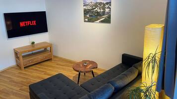 A modern living room with a large flat-screen TV displaying 'NETFLIX', a wooden coffee table, a dark sofa, a small round table with a plant, and a large floor lamp.
