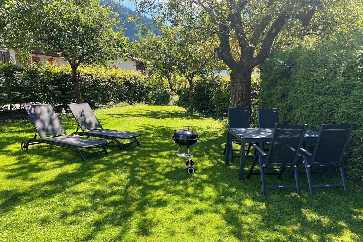 A grassy outdoor area with a barbecue grill, lounge chairs, and a dining table with chairs.