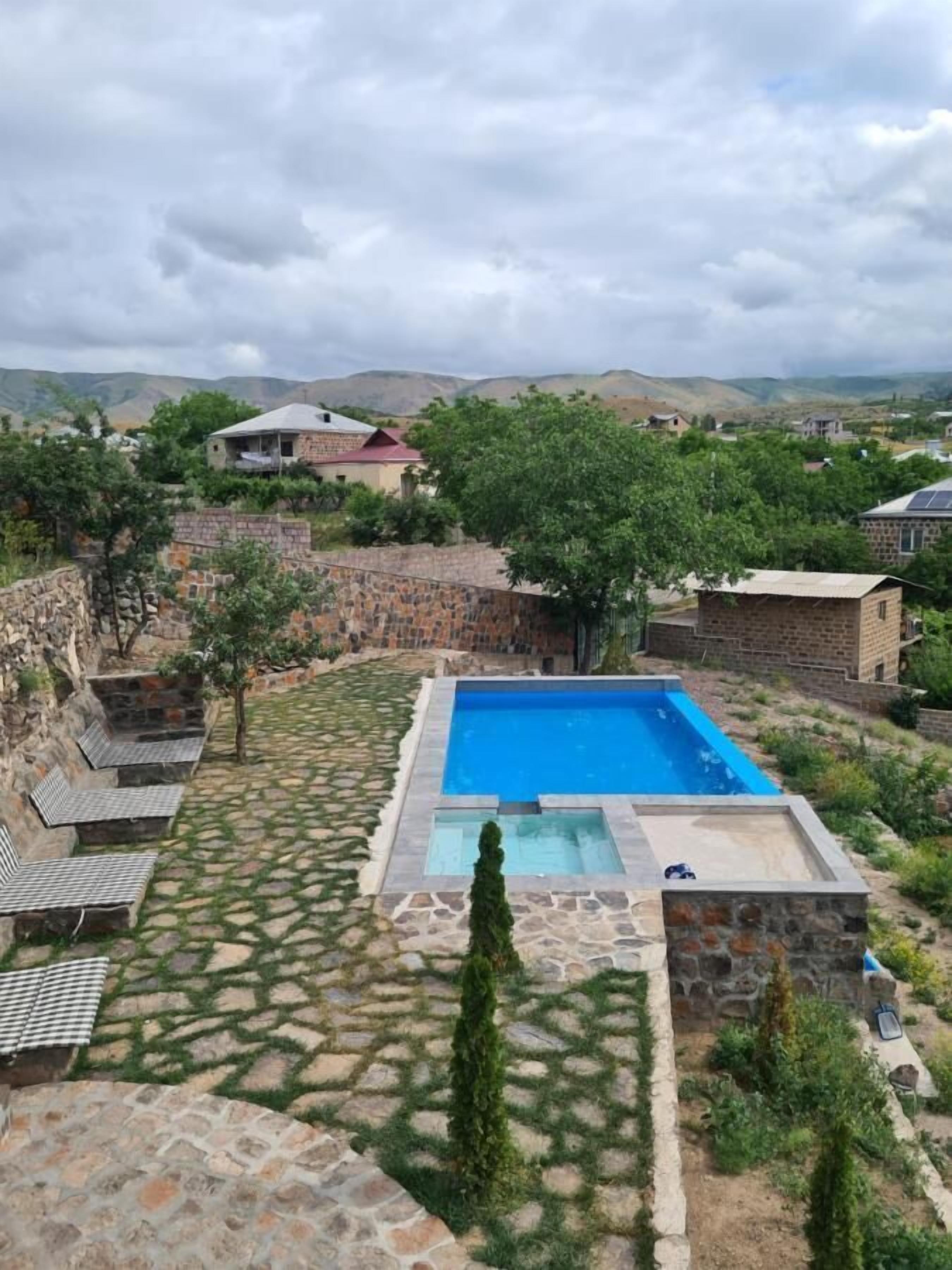 A rectangular swimming pool with a stone border, surrounded by a stone patio with seating areas and a view of a hilly landscape in the background.