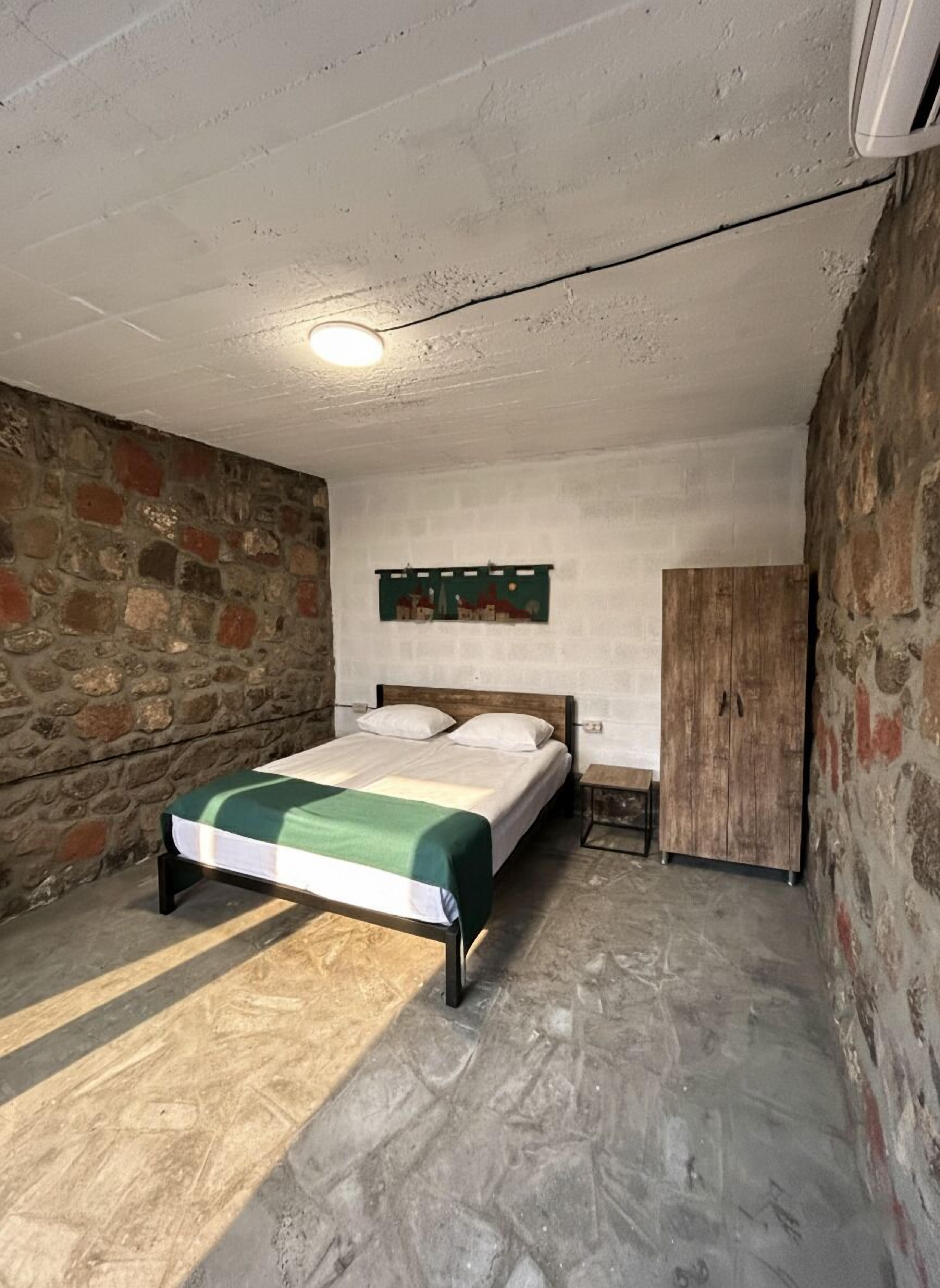 Een slaapkamer met een bed, een nachtkastje, een houten garderobe, een stenen muur en een plafondlamp.