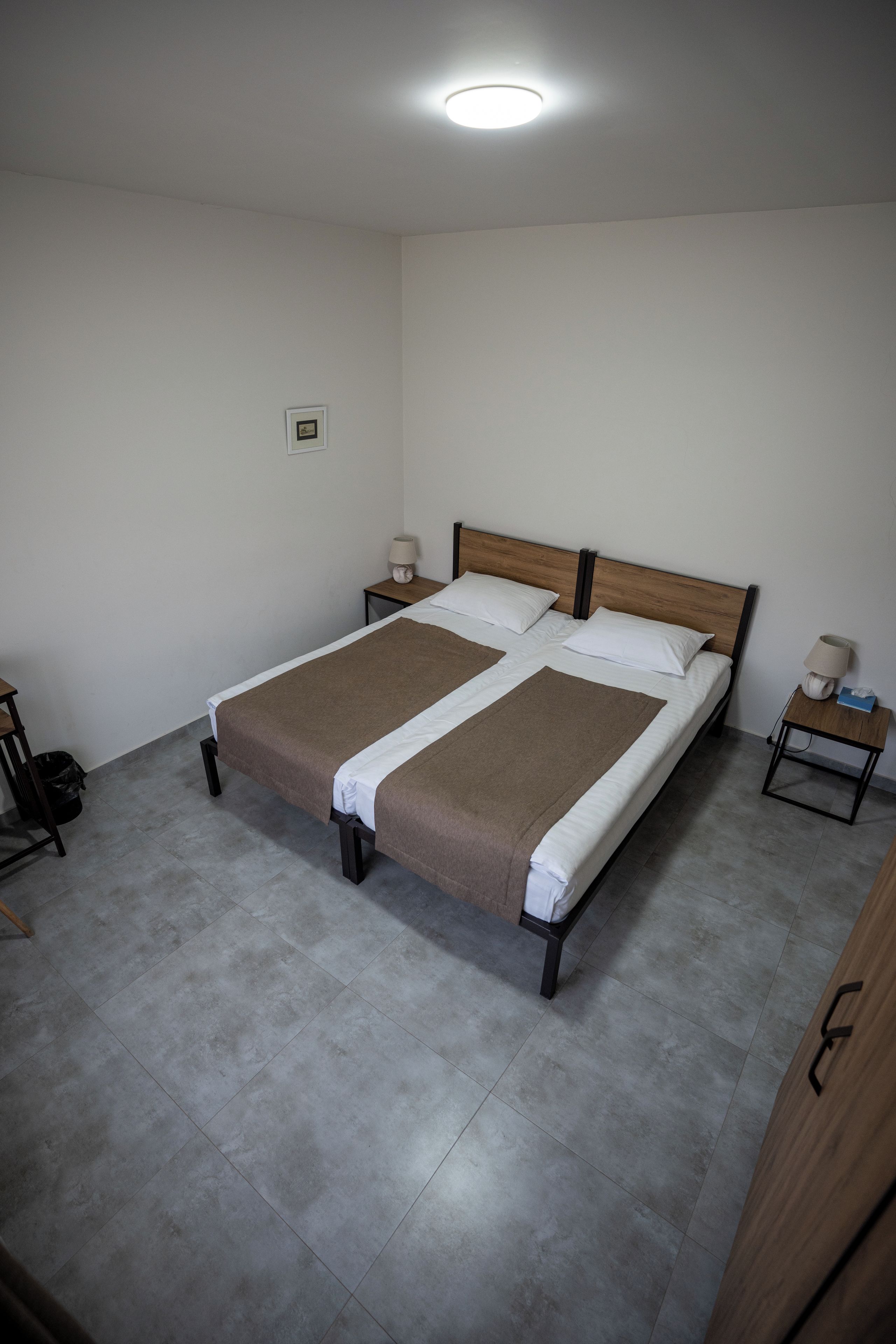 A bedroom with a large bed, two bedside tables, a ceiling light, and a small trash bin.