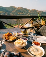 Mesa servida con diversos alimentos y una copa de vino, con vistas a un paisaje montañoso.