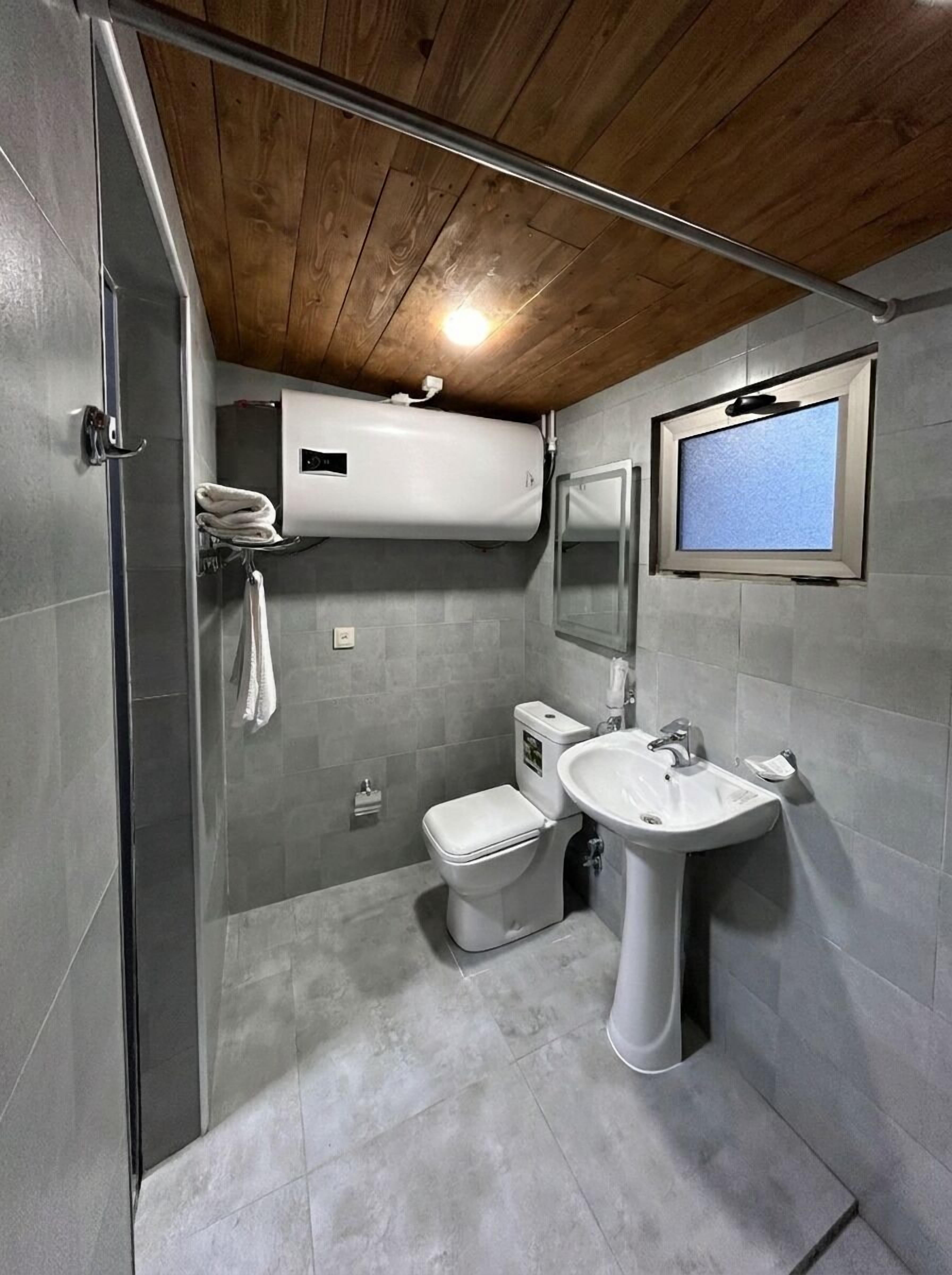 Un baño moderno con ducha, inodoro y lavabo, con baldosas grises y vigas de madera en el techo.