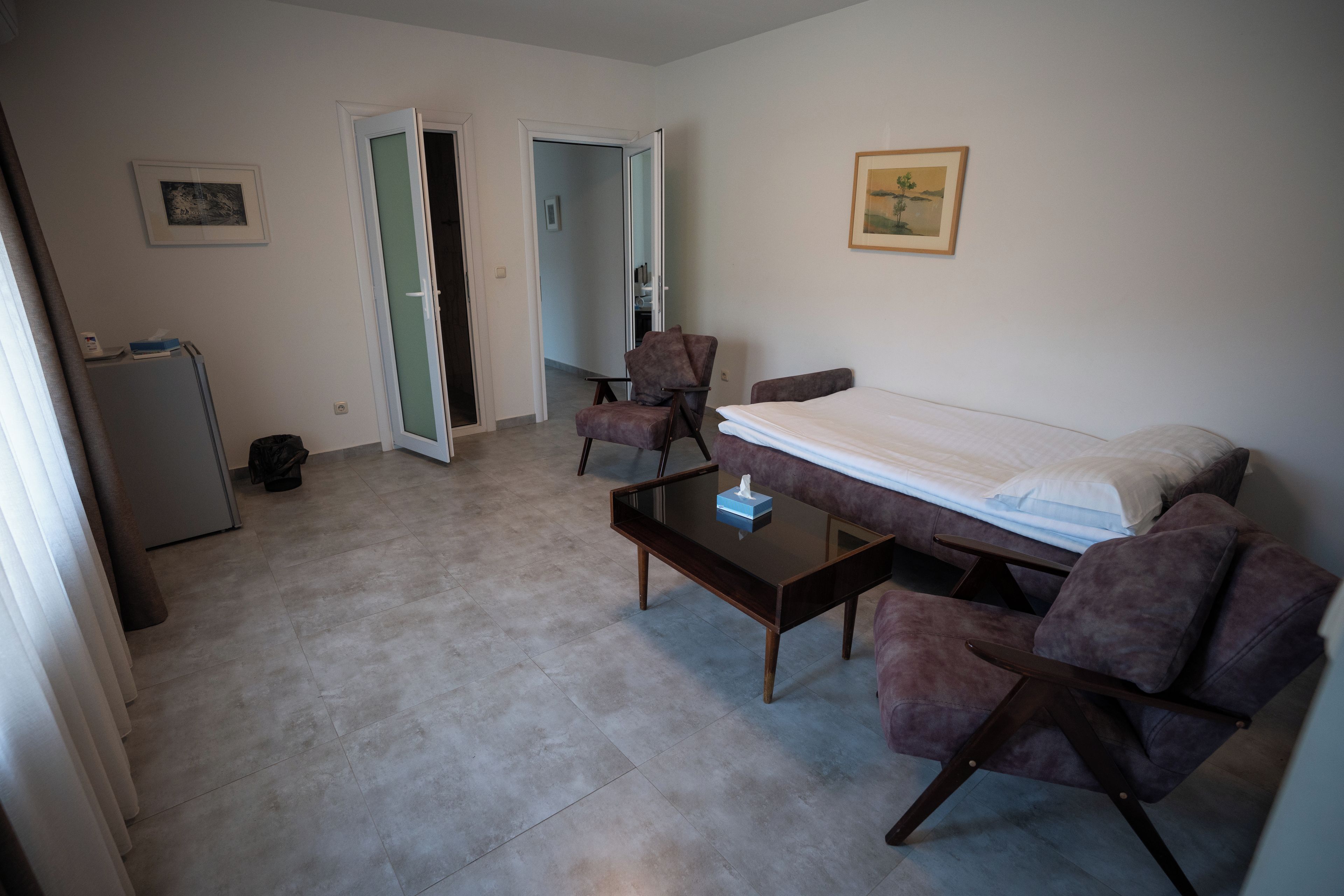 A compact room with a bed, a desk, two chairs, a small table, and a refrigerator.
