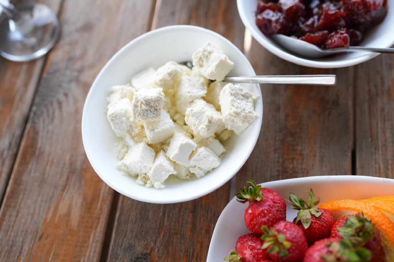 Un tazón de queso fresco con una cuchara, fresas y un tazón de compota de frutas sobre una mesa de madera.