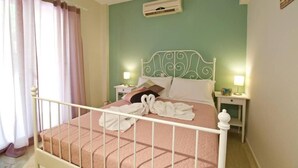 A neatly made bed with a pink bedspread and white pillows, a white metal headboard, and an air conditioning unit on the wall. - Blue & Green Apts - Dassia, Corfu, 1 (Dassia)