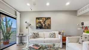 Ein modernes Wohnzimmer mit einem Glastisch, weißen Sofas, einem großen Stadtbild und Blick auf eine Wüstenlandschaft durch eine Schiebetür.