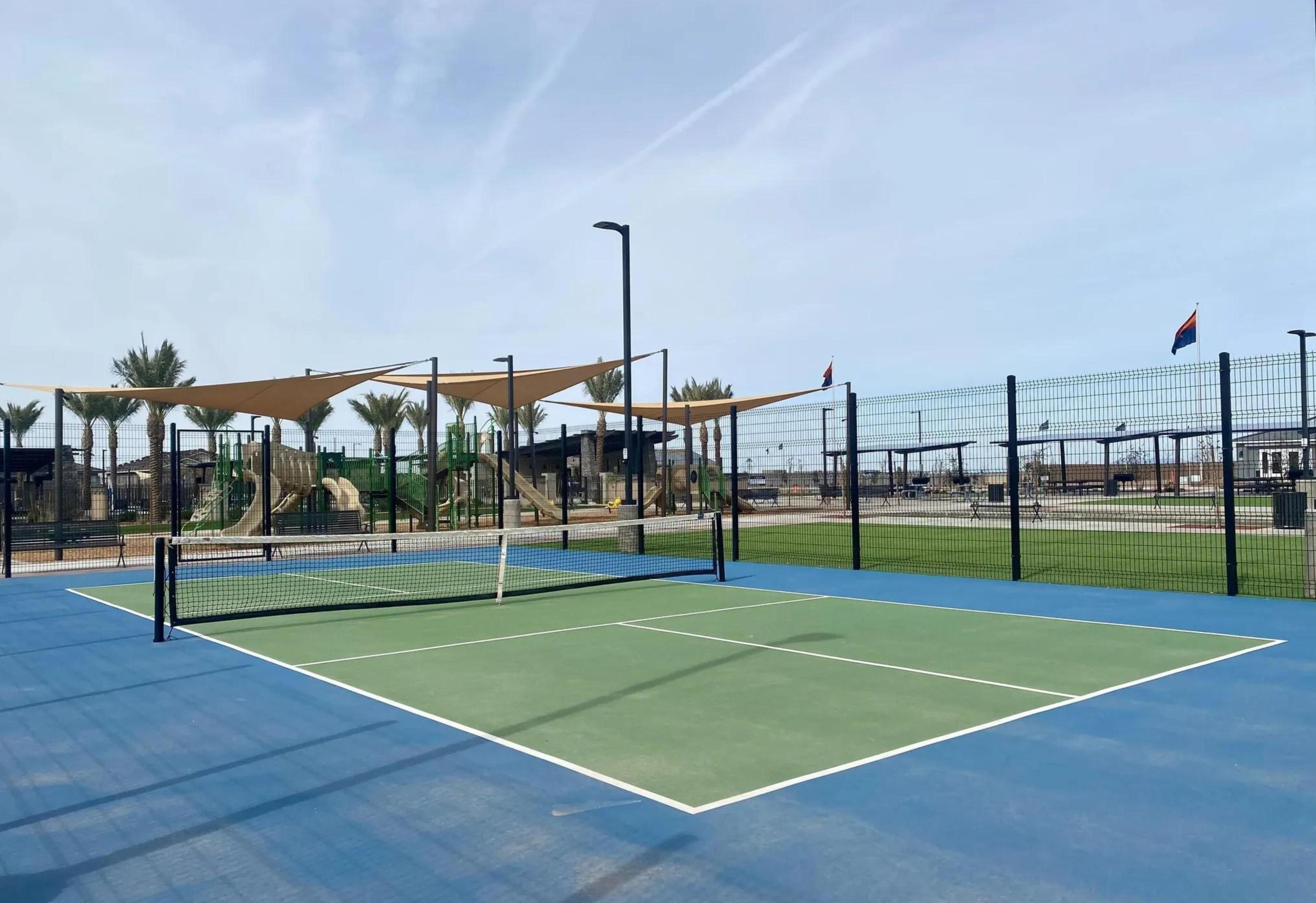 An outdoor tennis court with a net, surrounded by a fence and palm trees.