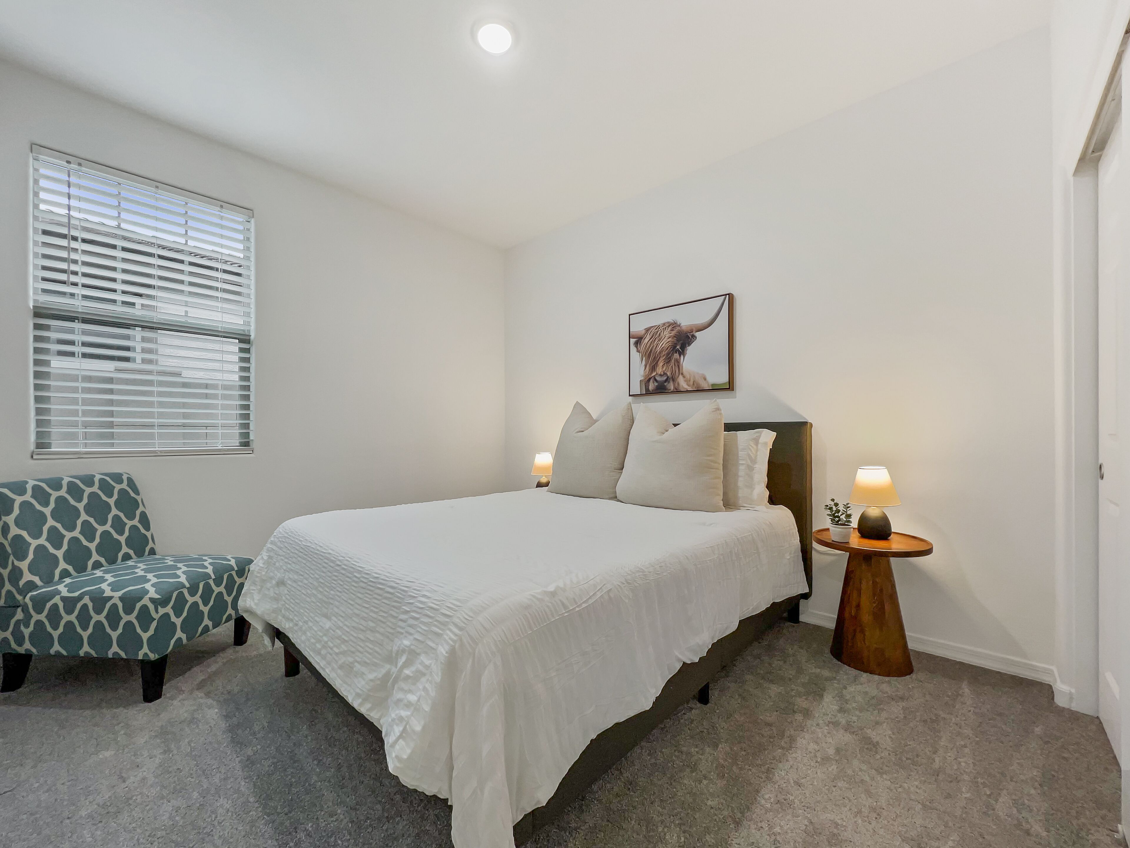 A bedroom with a bed, a chair, a bedside table with a lamp, a window with blinds, and a picture on the wall.