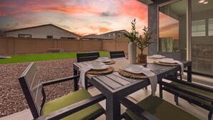 An outdoor dining area with a table set for four, surrounded by chairs, and a view of a sunset over a residential area. - 3BR w playroom, community pool/spa! (Queen Creek)