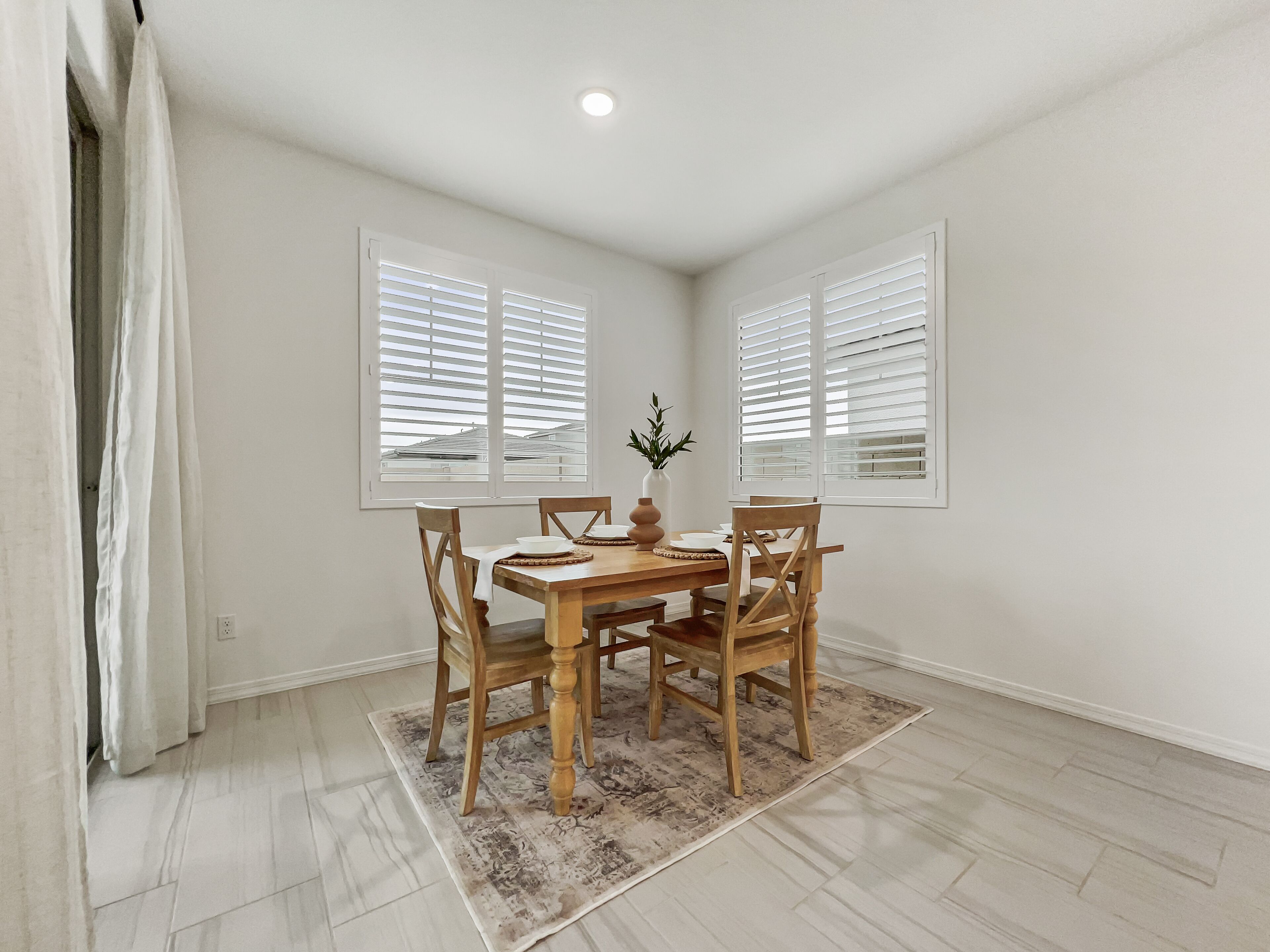 A dining area with a wooden table, four chairs, a vase with a plant, and two windows with white blinds.