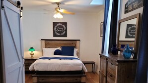 Une chambre avec un plancher en bois, un lit avec une literie blanche et bleue, un ventilateur de plafond, une table de chevet avec une lampe et une commode agrémentée d’objets décoratifs.